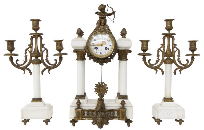 Een drie-delig witmarmeren klokkenstel bestaande uit een pendule en twee kandelaren. Versierd met amor figuurtjes.