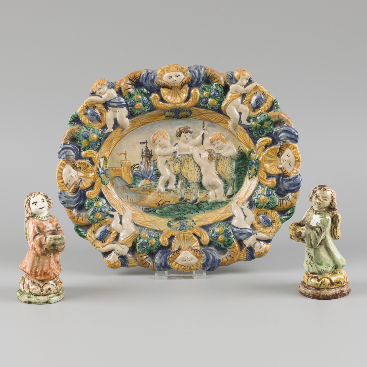 Een majolica schotel met voorstelling van putti die spelen met een geit, daarbij twee kaarsenhoudertjes, 20e eeuw.